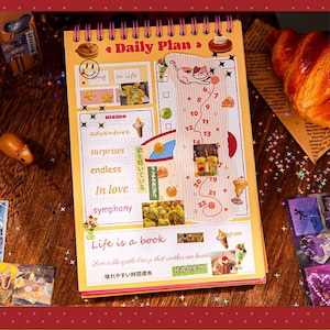 May include: A spiral-bound daily planner with a yellow cover featuring the text "Daily Plan" and various illustrations. The planner includes sections for notes and a numbered list. A croissant and stickers are also visible.
