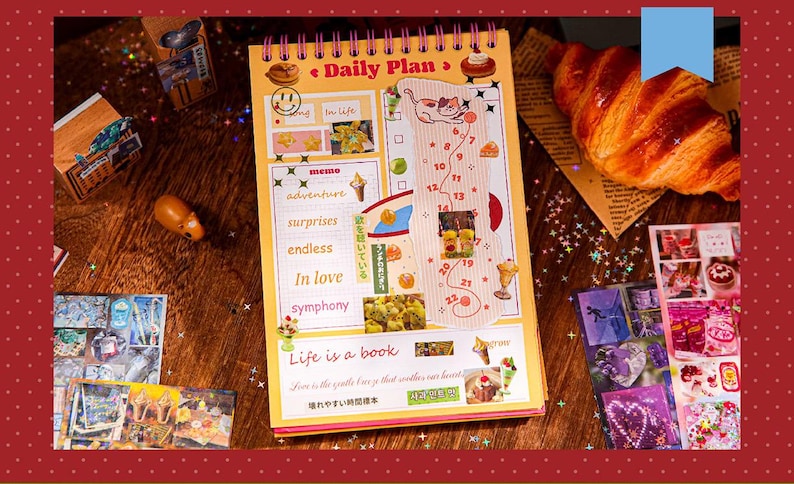 May include: A spiral-bound daily planner with a yellow cover featuring the text "Daily Plan" and various illustrations. The planner includes sections for notes and a numbered list. A croissant and stickers are also visible.