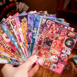 May include: A hand holding a fan of colorful stickers with various designs. The stickers feature images of food, flowers, and other decorative elements. The stickers are in various colors, including pink, red, blue, and green.