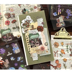 May include: A collection of decorative stickers featuring floral and botanical designs. The stickers include images of flowers, storefronts, and vintage-style elements, arranged on a notebook and clipboard.