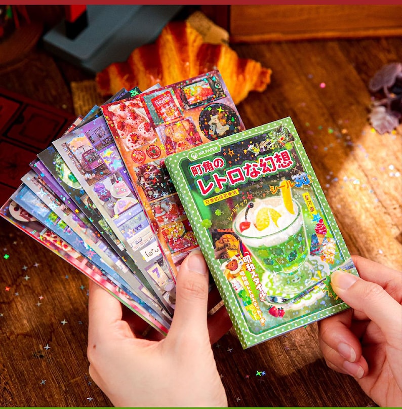 May include: A collection of colorful stickers and a green book with Japanese text are held in a person's hands. The stickers feature various designs, including food and scenes. A croissant is in the background.
