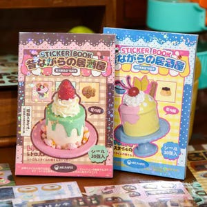 Op de afbeelding: Twee stickerboeken met Japanse tekst en dessertillustraties. Eén boek toont een cake met aardbei, de andere een cupcake. Beide boeken hebben roze en blauwe ontwerpen. De tekst bevat "Sticker Book".