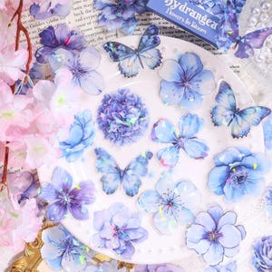 Puede incluir: Una colección de pegatinas de flores y mariposas azules y moradas dispuestas sobre un plato blanco. Las pegatinas varían en tamaño y forma, con algunos detalles iridiscentes. La imagen incluye el texto "Hydrangea Flowers In Bloom".