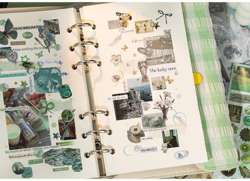 May include: A six-ring binder planner open to two pages filled with various stickers, including a butterfly, Snoopy, flowers, and text snippets like "The lucky ones" and "Keep smiling life is beautiful." The pages feature a collage of green-toned images.