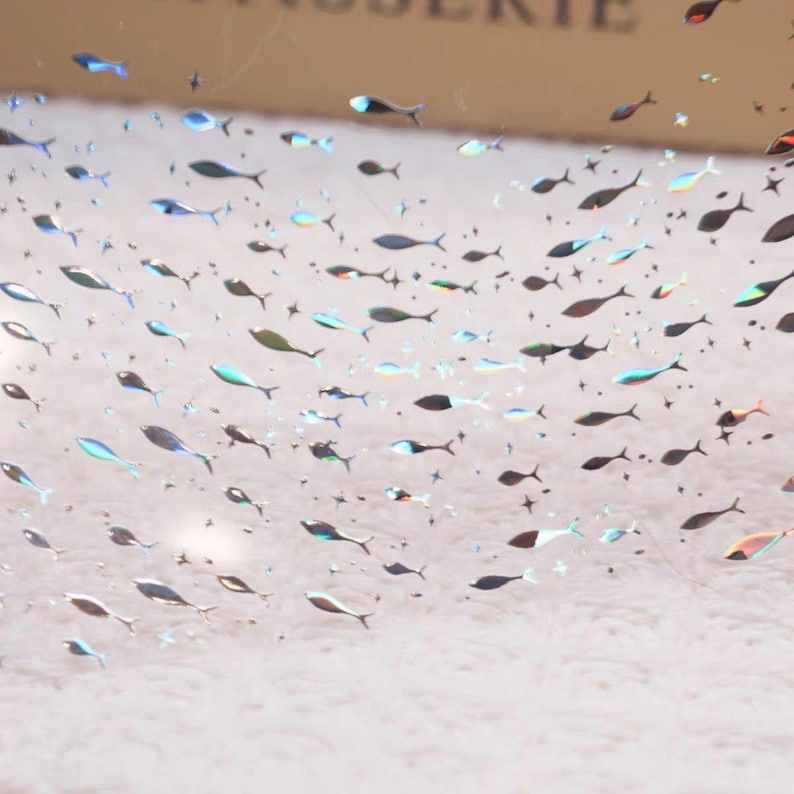 May include: A collection of small, iridescent fish-shaped stickers scattered across a light-colored surface. The stickers vary in color, including blue, green, and black, and appear to be reflective. The overall effect is a shimmering, aquatic theme.