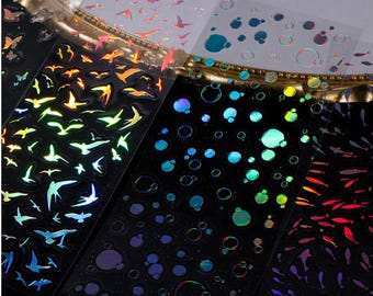 May include: A collection of iridescent stickers featuring various designs. The stickers include images of birds in flight, bubbles, and fish. The stickers have a holographic effect, with colors shifting from blue to green to orange.