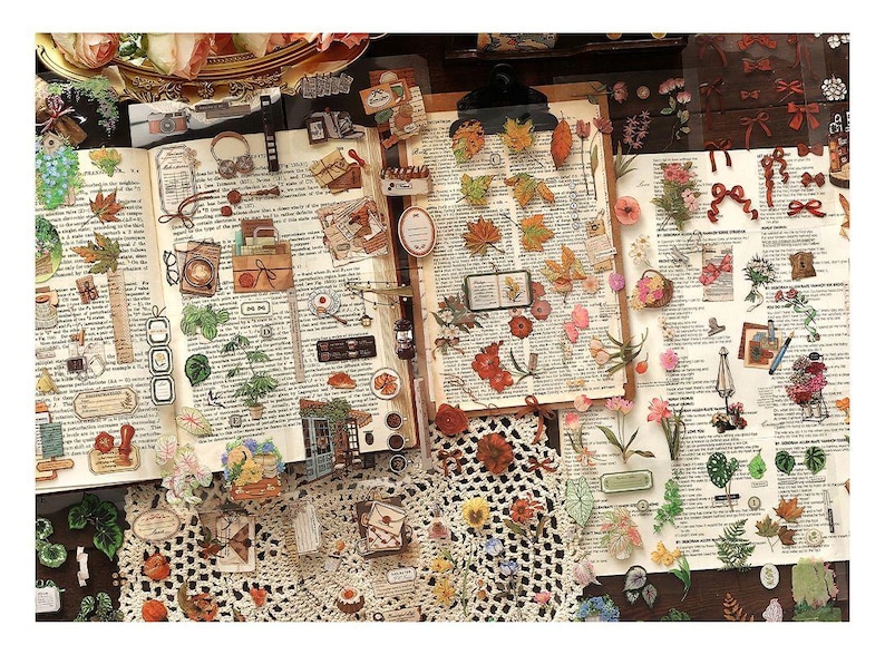 May include: An open book with vintage-style stickers and embellishments. The stickers include leaves, flowers, bows, and small illustrations. The background is a printed page with text. A white crocheted doily is in the foreground.