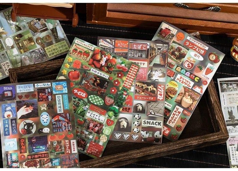 Op de afbeelding: Een verzameling decoratieve stickers met Japanse thema's. De stickers tonen afbeeldingen van eten, dieren en Japanse tekst. De stickers zijn gerangschikt op transparante vellen, met een verscheidenheid aan kleuren en vormen.