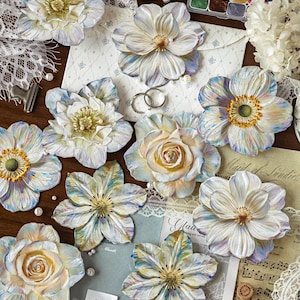 Pode incluir: Uma coleção de ornamentos florais decorativos com pétalas pintadas em branco, azul e amarelo com superfícies texturizadas. As flores estão dispostas ao lado de um conjunto de aguarelas, renda branca e papelaria com o texto This package is happy to see you too.
