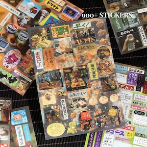 Op de afbeelding: Vellen decoratieve stickers met diverse ontwerpen, waaronder stripfiguren, voedsel en Japanse tekst. De stickers zijn kleurrijk en hebben een verscheidenheid aan vormen en maten. De tekst "900+ STICKERS" is zichtbaar.
