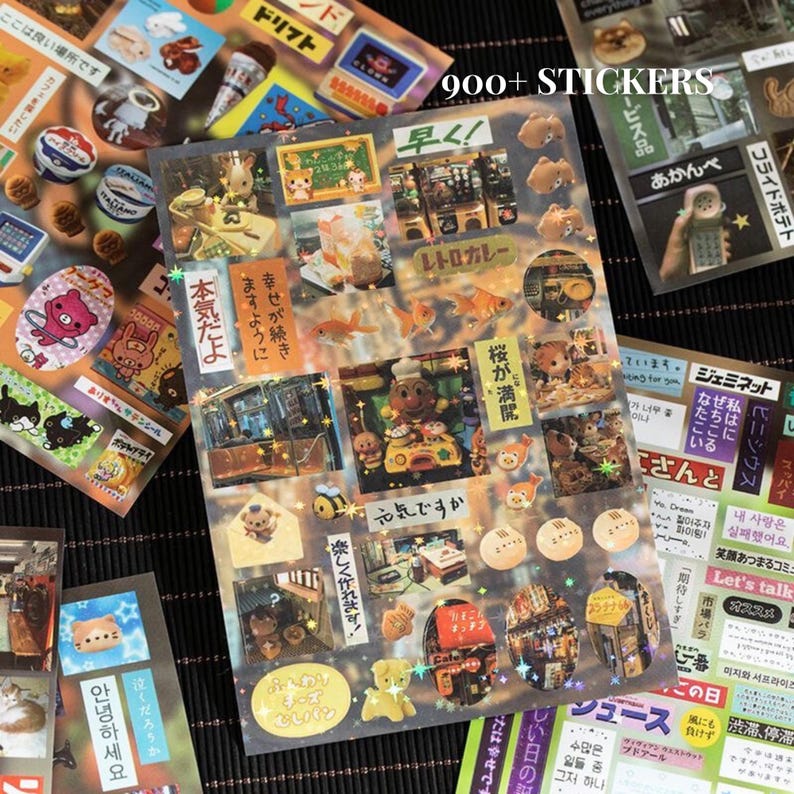 Op de afbeelding: Vellen decoratieve stickers met diverse ontwerpen, waaronder stripfiguren, voedsel en Japanse tekst. De stickers zijn kleurrijk en hebben een verscheidenheid aan vormen en maten. De tekst "900+ STICKERS" is zichtbaar.