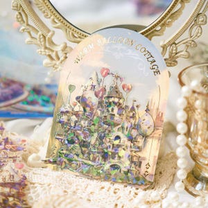 Op de afbeelding: Een holografische sticker met een fantasie kasteelontwerp, compleet met hartvormige ballonnen. De sticker is voorzien van de tekst "Warm Balloon Cottage" in goud. Het kasteel is gedetailleerd en het kleurenpalet is zacht en pastel.