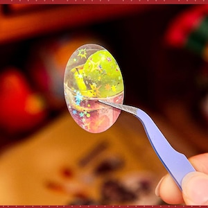May include: An oval-shaped sticker with a clear, glittery background, featuring a green object and star-shaped accents. The sticker is held by blue tweezers, with a blurred background of other objects.