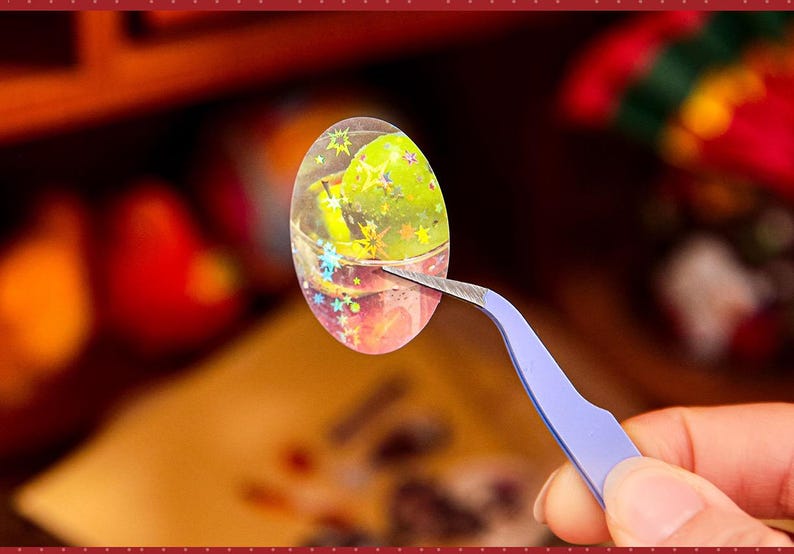 May include: An oval-shaped sticker with a clear, glittery background, featuring a green object and star-shaped accents. The sticker is held by blue tweezers, with a blurred background of other objects.
