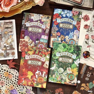 May include: Four sticker books with decorative covers. Each book features the text "STICKER BOOK" and "200" on the cover. The covers have different designs, including floral, botanical, and vintage illustrations. The books are arranged on a wooden surface.