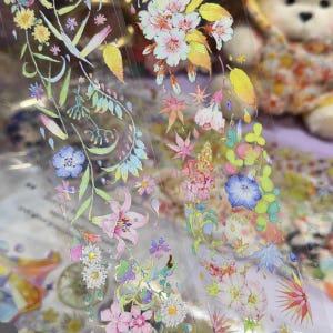 May include: A roll of clear adhesive tape featuring a repeating pattern of colorful botanical illustrations, including pink lilies, blue morning glories, white daisies, and various green leaves with a subtle holographic finish throughout.