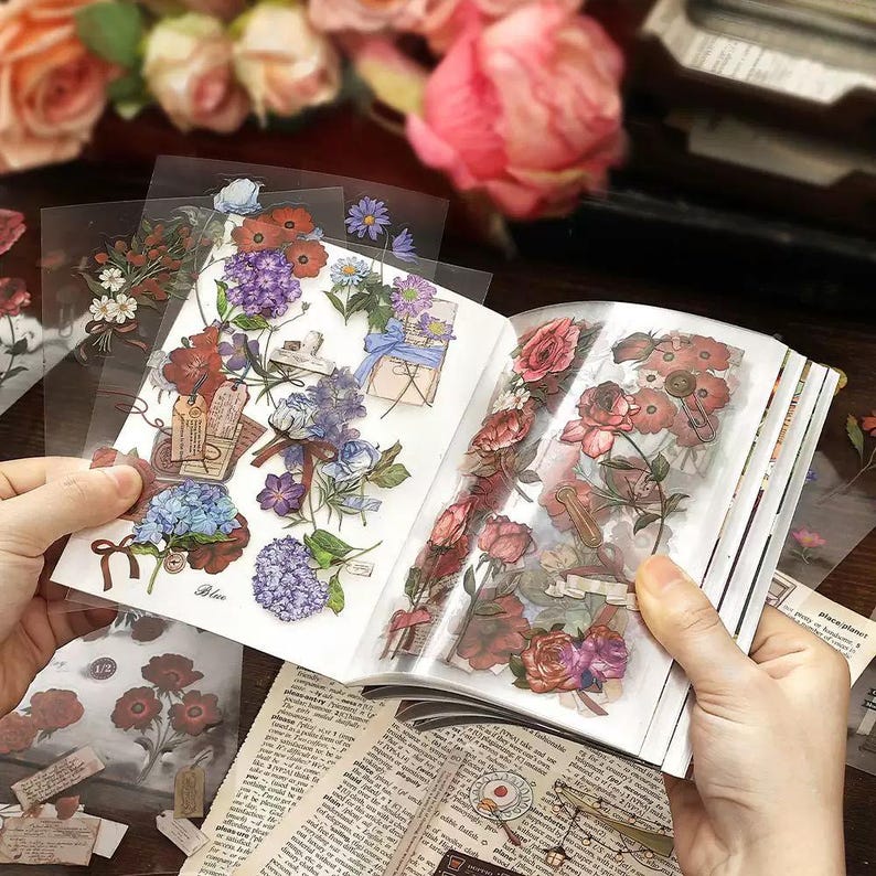 May include: A journal open to pages of floral stickers, featuring roses, hydrangeas, and other flowers in shades of red, purple, and blue. The stickers are on clear backing, with some loose stickers visible. The background includes blurred flowers.