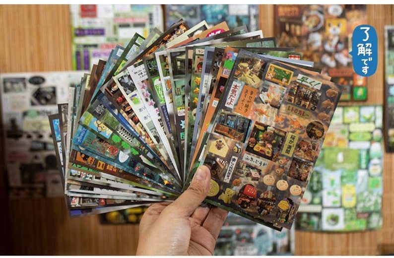 Op de afbeelding: Een hand houdt een waaier van kleurrijke stickers vast. De stickers tonen diverse ontwerpen, waaronder Japanse tekst, voedselillustraties en landschapsgezichten. De stickers zijn in verschillende maten en vormen, met een glanzende afwerking.