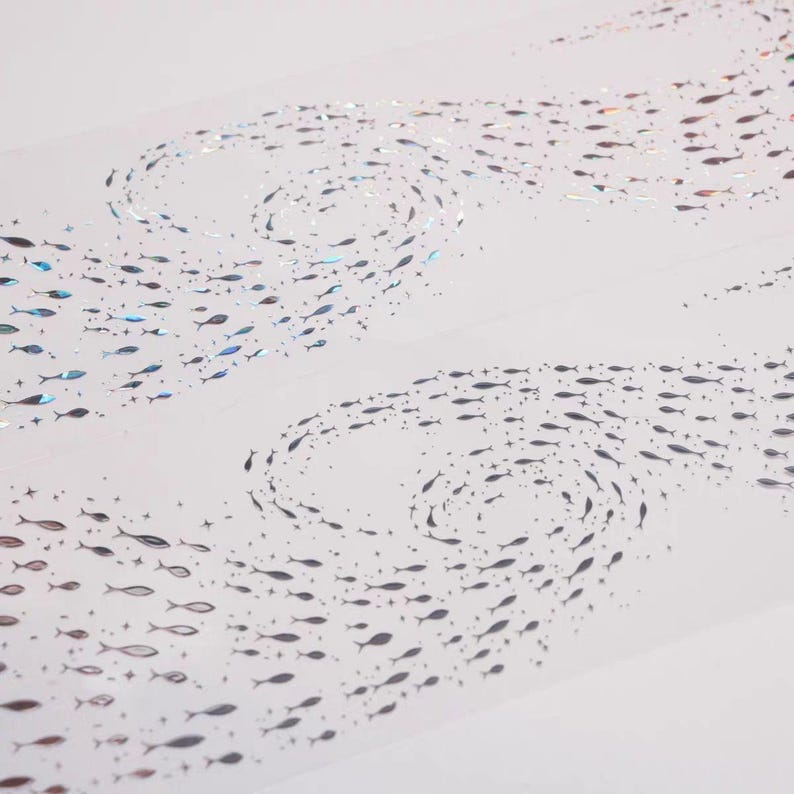 May include: Decorative tape featuring a repeating pattern of small fish in swirling formations. The fish are primarily silver with iridescent accents, creating a shimmering effect against the white background. The design evokes a sense of movement and the ocean.