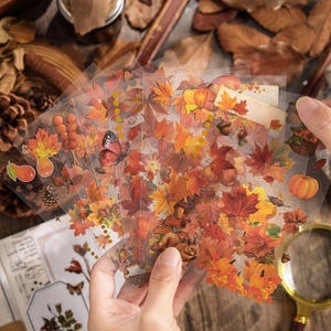 May include: Sheets of clear stickers with autumn-themed illustrations. The stickers depict colorful maple leaves, pumpkins, acorns, butterflies, and pears in shades of orange, red, and yellow. The stickers are held in a person's hand.