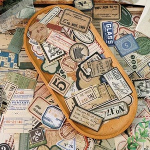 Könnte beinhalten: Eine Holzschale gefüllt mit Papier-Ephemera im Vintage-Stil, darunter Etiketten, Tickets und Stempel. Die Artikel weisen verschiedene Schriftarten, Farben und Designs auf, mit Texten wie "Lancaster, N.H." und "Vanilla Chocolate".