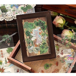 May include: A framed botanical collage featuring green leaves and a vintage paper background. Surrounding the frame are various floral and plant-themed stickers and embellishments, with a wooden box and roses in the background.