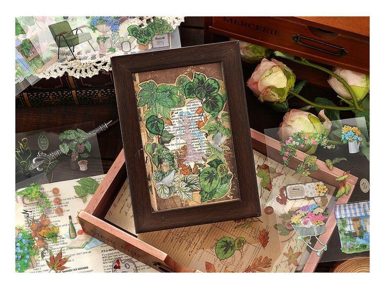 May include: A framed botanical collage featuring green leaves and a vintage paper background. Surrounding the frame are various floral and plant-themed stickers and embellishments, with a wooden box and roses in the background.
