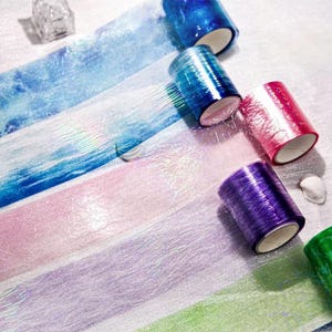 Puede incluir: Una variedad de rollos de cinta washi colorida en tonos de azul, rosa, morado y verde. La cinta tiene un acabado brillante e iridiscente, y está desenrollada para mostrar los patrones decorativos. Un pequeño recipiente transparente y facetado está en el fondo.
