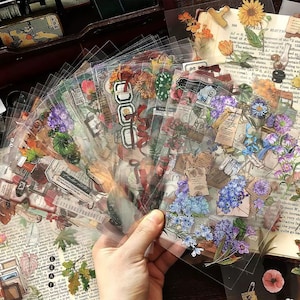 May include: A hand holds a fan of clear stickers with floral and vintage-themed designs. The stickers feature illustrations of flowers, leaves, and old documents. Colors include shades of purple, blue, red, and brown.