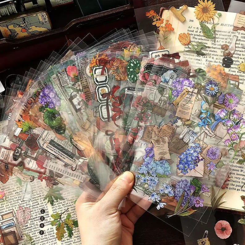 May include: A hand holds a fan of clear stickers with floral and vintage-themed designs. The stickers feature illustrations of flowers, leaves, and old documents. Colors include shades of purple, blue, red, and brown.