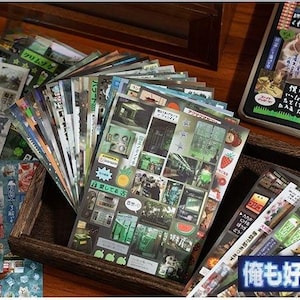 Op de afbeelding: Een verzameling decoratieve stickers en briefpapier, waaronder vellen met Japanse tekst en kleurrijke illustraties. De stickers bevatten verschillende ontwerpen, waaronder afbeeldingen van voedsel, gebouwen en tekst. De items zijn gerangschikt in een houten doos.