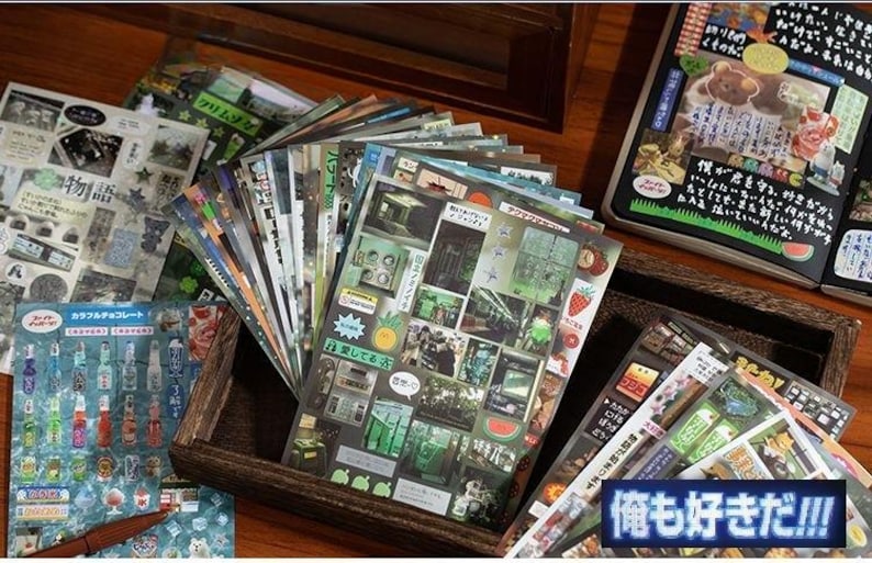Op de afbeelding: Een verzameling decoratieve stickers en briefpapier, waaronder vellen met Japanse tekst en kleurrijke illustraties. De stickers bevatten verschillende ontwerpen, waaronder afbeeldingen van voedsel, gebouwen en tekst. De items zijn gerangschikt in een houten doos.