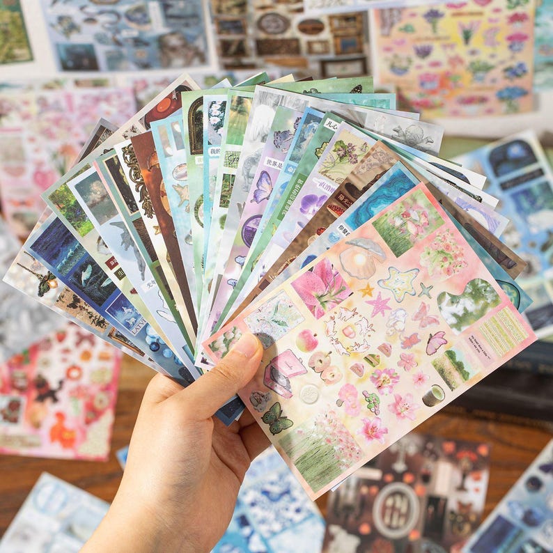 Hand holding multiple decorative collage sticker sheets showing a variety of colors, themes, and illustration styles for creative paper crafts.