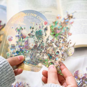 Op de afbeelding: Een holografische sticker met een grillig kasteelontwerp met hartvormige ballonnen. De sticker heeft een gouden rand en de tekst "Balloon Cottage". Het kasteel heeft meerdere torens en is versierd met pastelkleuren.