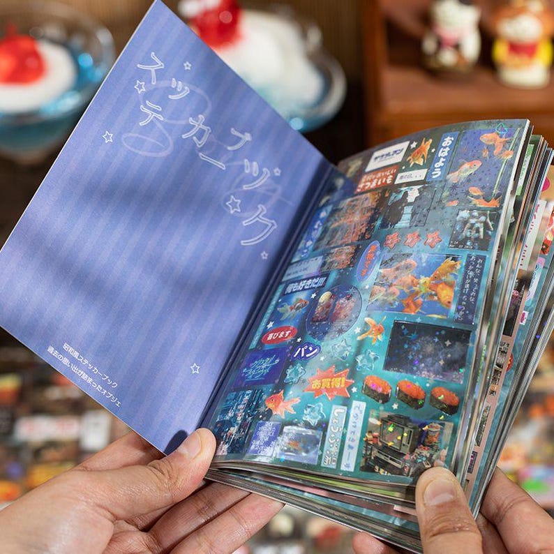 Op de afbeelding: Een open stickerboek met een blauwe kaft en pagina's vol kleurrijke stickers. De kaft heeft Japanse tekst en sterillustraties. De stickers bevatten verschillende afbeeldingen, waaronder vissen, sushi en Japanse tekst.