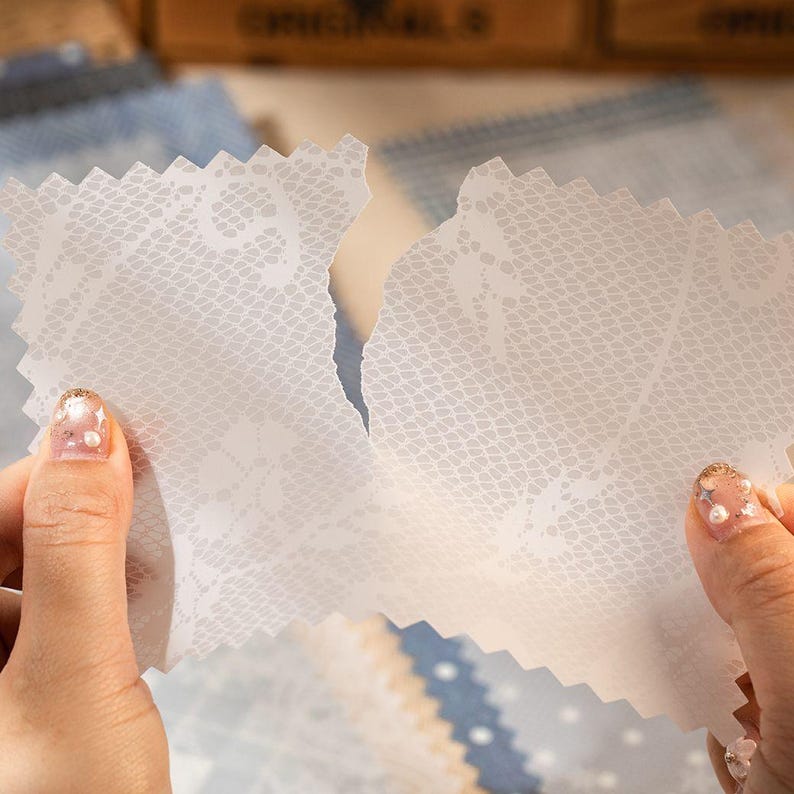 May include: Two pieces of white lace-patterned paper are being torn apart, revealing a delicate floral design. The paper has a scalloped edge and is held by hands with manicured nails. Other patterned papers are in the background.