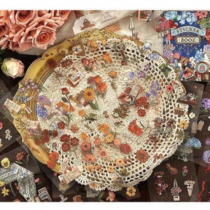 May include: A collection of floral stickers and a sticker book are arranged on a table. The stickers feature various flowers, books, and other decorative elements. A gold-rimmed tray with a doily and roses are also present.
