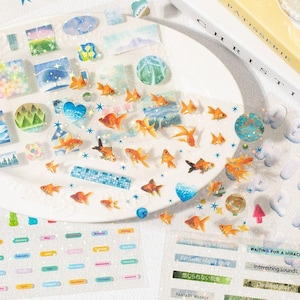 Op de afbeelding: Decoratieve stickers met goudvissen, landschappen en tekst. De stickers hebben verschillende vormen, waaronder vierkanten, cirkels en rechthoeken, met oranje, blauwe en groene tinten. Sommige stickers hebben tekst zoals "Fantastic reality story" en "FANTASY WEEKLY."
