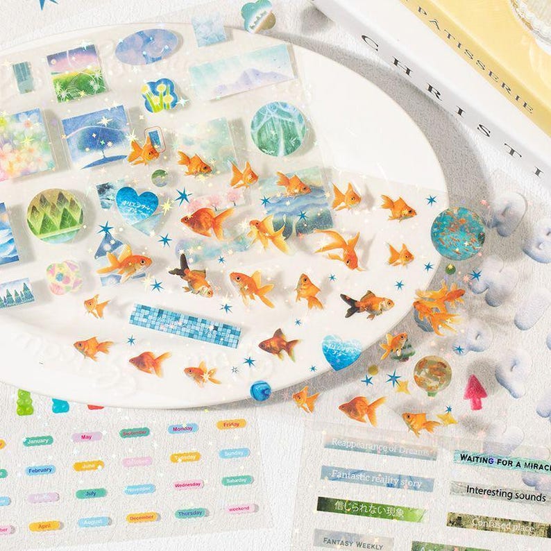 Op de afbeelding: Decoratieve stickers met goudvissen, landschappen en tekst. De stickers hebben verschillende vormen, waaronder vierkanten, cirkels en rechthoeken, met oranje, blauwe en groene tinten. Sommige stickers hebben tekst zoals "Fantastic reality story" en "FANTASY WEEKLY."
