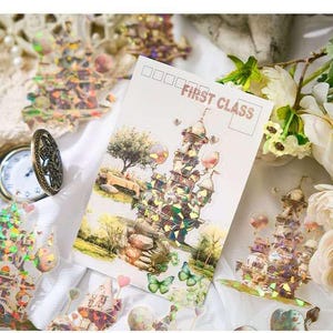 Op de afbeelding: Een ansichtkaart met een kasteelillustratie en de tekst "FIRST CLASS". De afbeelding toont een grillig kasteelontwerp met holografische accenten, vlinders en een boom. Decoratieve stickers en een zakhorloge zijn ook zichtbaar.