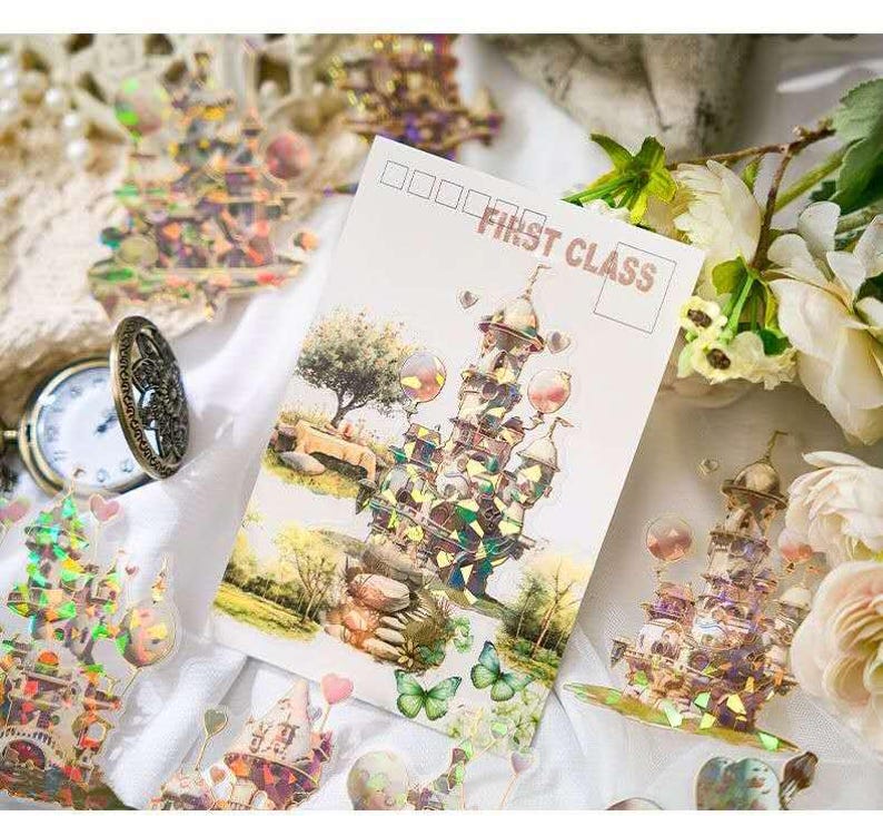 Op de afbeelding: Een ansichtkaart met een kasteelillustratie en de tekst "FIRST CLASS". De afbeelding toont een grillig kasteelontwerp met holografische accenten, vlinders en een boom. Decoratieve stickers en een zakhorloge zijn ook zichtbaar.