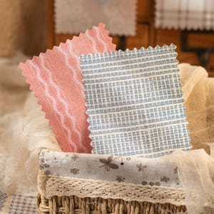 May include: Two rectangular fabric swatches with pinked edges. One swatch is coral with white vertical wavy lines. The other swatch is light blue with a white grid pattern. The swatches are resting inside a woven basket lined with floral fabric.