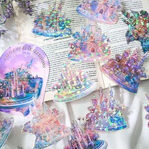 Op de afbeelding: Een verzameling iriserende stickers met kasteelontwerpen in roze, blauwe en paarse tinten. E&eacute;n sticker leest "Castle of Pink Illusions". De stickers zijn gerangschikt op een pagina van een boek en een wit oppervlak.