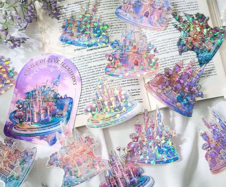 Op de afbeelding: Een verzameling iriserende stickers met kasteelontwerpen in roze, blauwe en paarse tinten. E&eacute;n sticker leest "Castle of Pink Illusions". De stickers zijn gerangschikt op een pagina van een boek en een wit oppervlak.