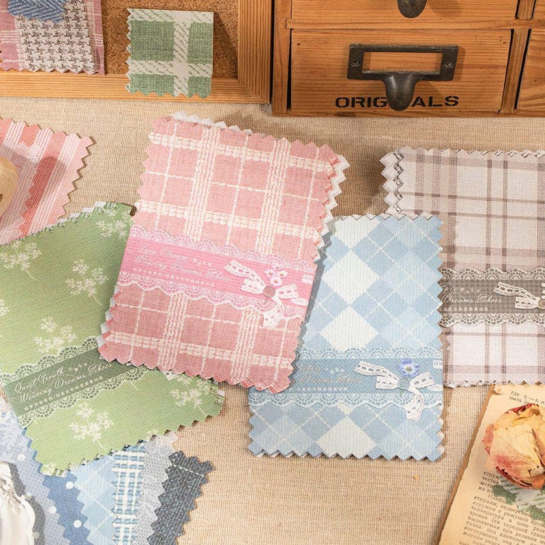 May include: A collection of rectangular fabric swatches with pink, green, blue, and beige plaid or floral patterns. Each swatch features a decorative lace band with a small bow and text, finished with pinked edges, arranged on a textured surface.