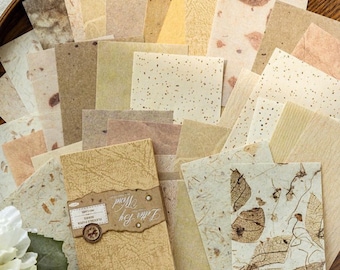 May include: A collection of handmade paper in various shades of beige, tan, and cream. Some sheets have leaf and speckle patterns. A small booklet with the text "Letter Joy Word" is included, along with a decorative band and a small, round clasp.