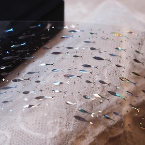 May include: A clear, iridescent sheet adorned with numerous small fish and star-shaped designs. The fish vary in color, displaying shades of blue, silver, and hints of other colors. The background is a soft, textured fabric.