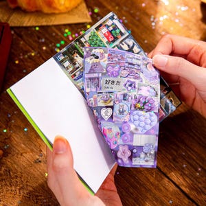 May include: A notebook with a green spine and white pages is held open, revealing a collage of purple and white stickers. The stickers feature various designs, including cartoon characters, bows, and floral patterns. Japanese text is visible on one of the stickers.