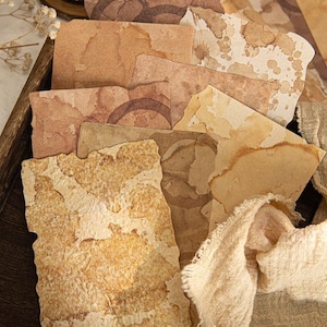 May include: A collection of rectangular paper sheets with uneven, deckled edges. The paper features various shades of brown, tan, and cream, with visible coffee-stain patterns, circular ring marks, and textured, fibrous surfaces in a layered arrangement.