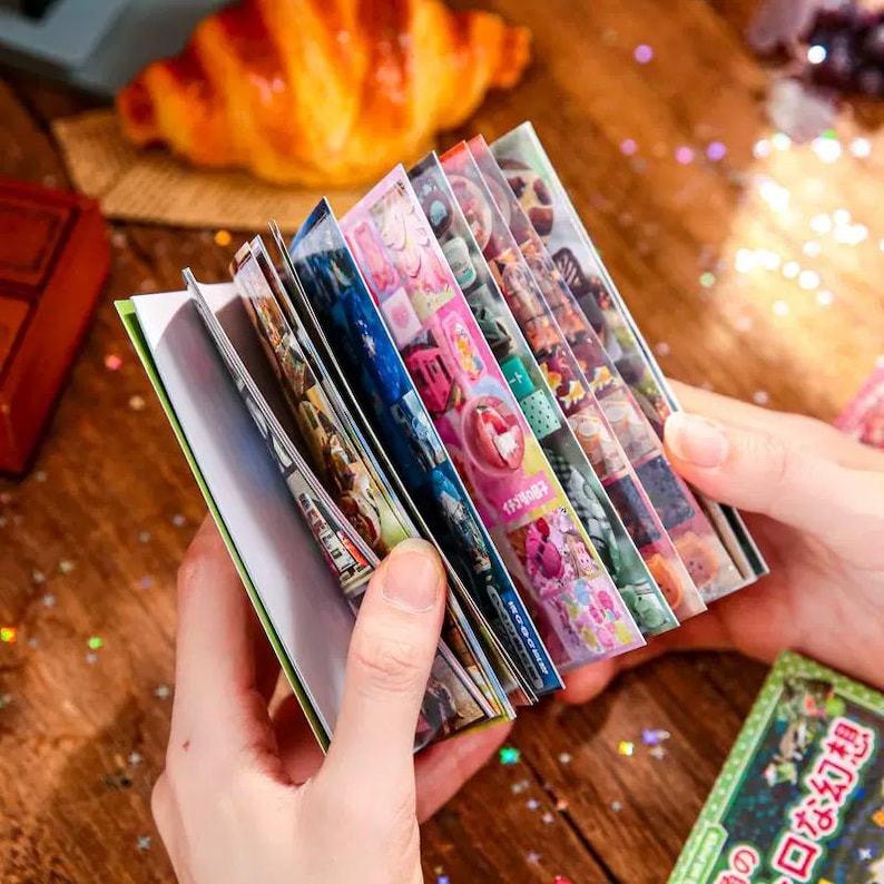 May include: A collection of colorful sticker sheets with various designs, held open to display the images. The stickers feature images of food, landscapes, and other decorative elements. The sheets are held in a person's hands.